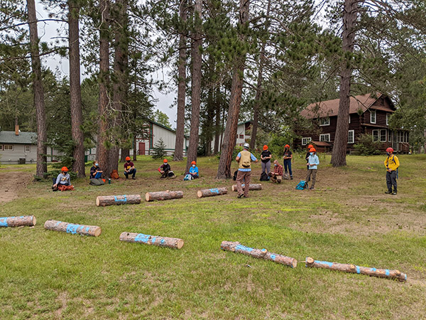 logs laid out on the ground with students lined up looking at them and listening to an instructor speak