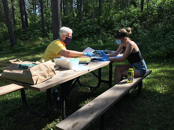 Two women with masks on sitting at a picnic table looking over old photos and documents