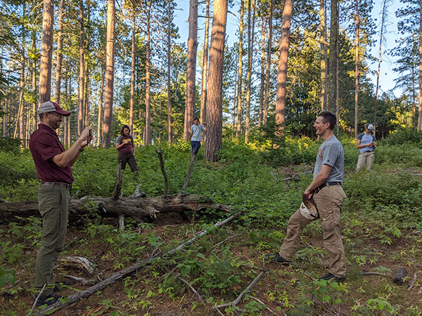 Eli Sagor holding a cell phone recording Kyle Gill in the forest with three onlookers in the background