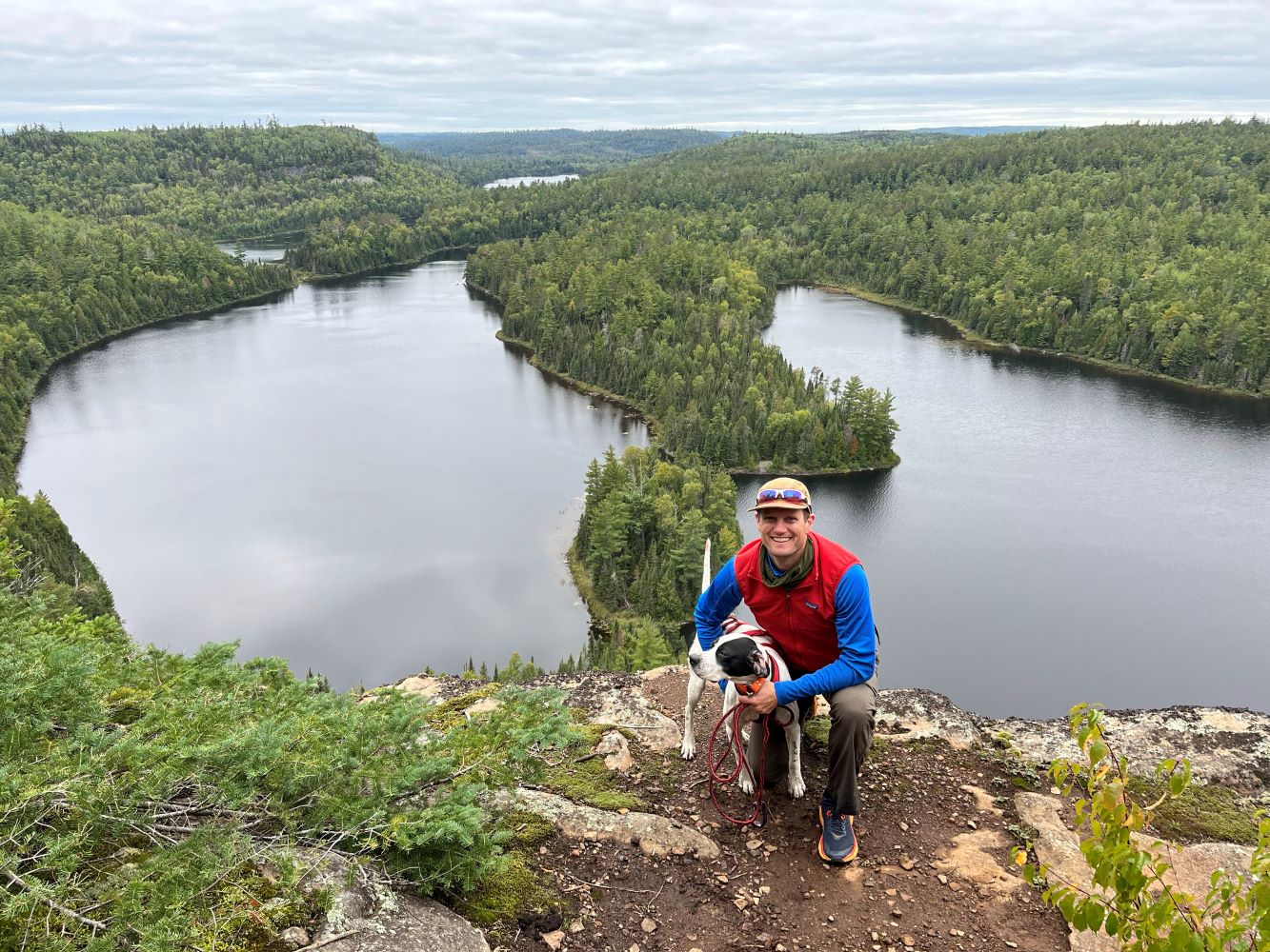 Kyle Gill kneels at the edge of a cliff holding his dog Arlo. In the background, a landscape of pines and lakes is seen past the cliff drop.