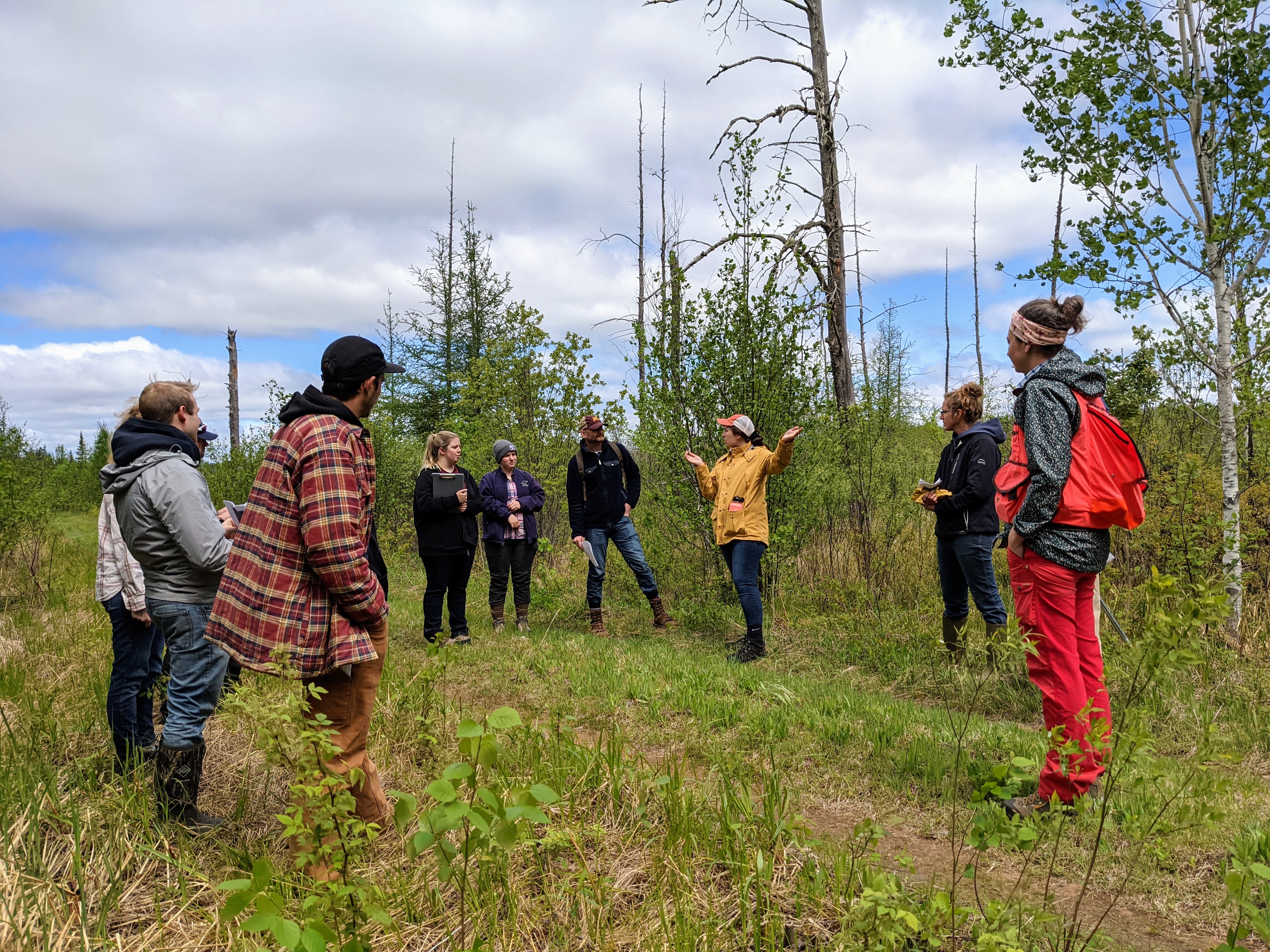 Several college-age students stand in a clear wooded area as their instructor speaks in the center.