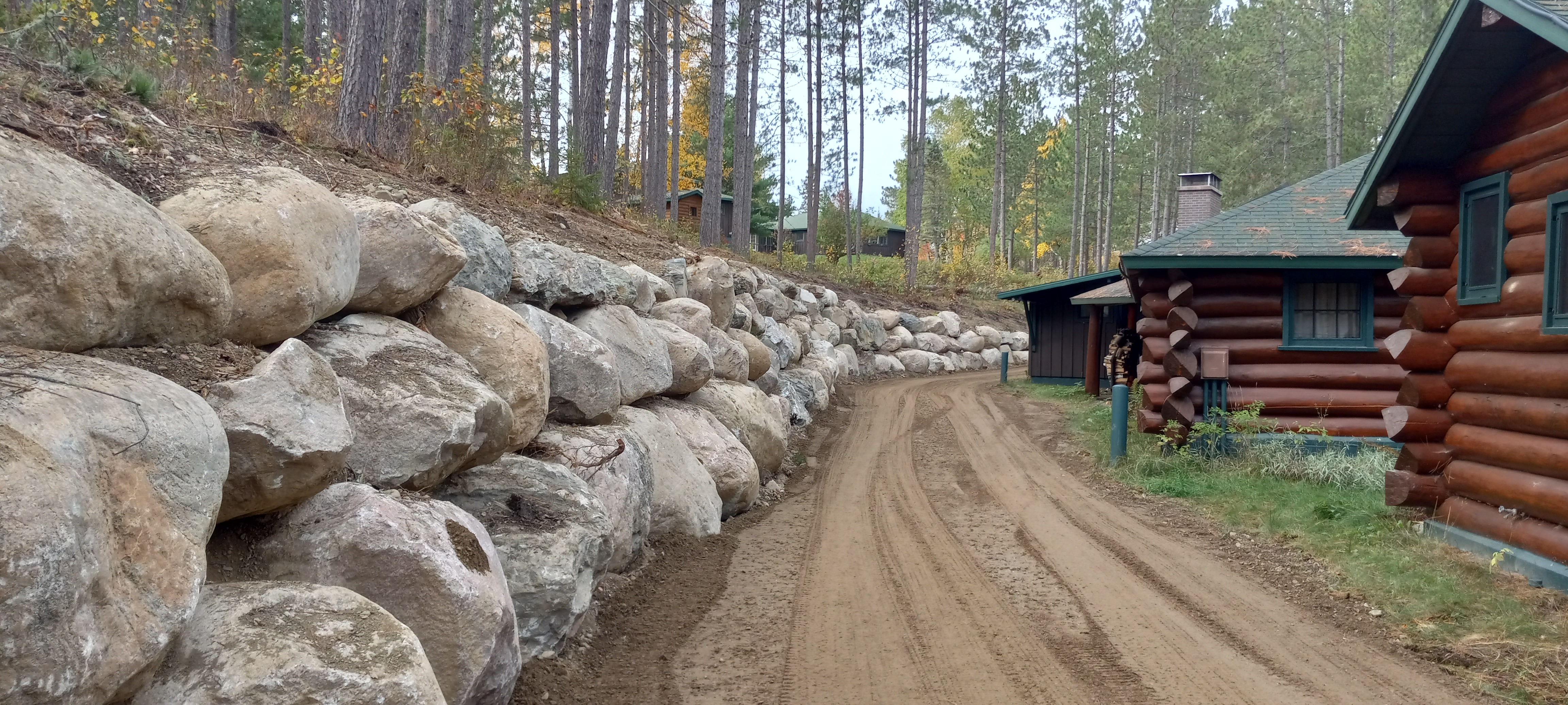 A dirt path stretches into the distance. On the right are log cabins, on the left are large boulders as part of the retaining wall.