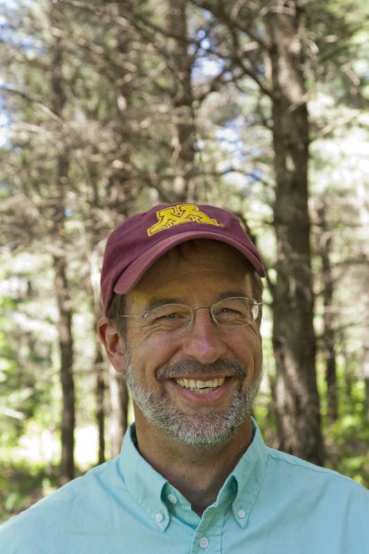 Andrew David, wearing a light blue button-up shirt and a maroon baseball cap with the gold block 'M' on the front, stands smiling in front of a stand of pines.