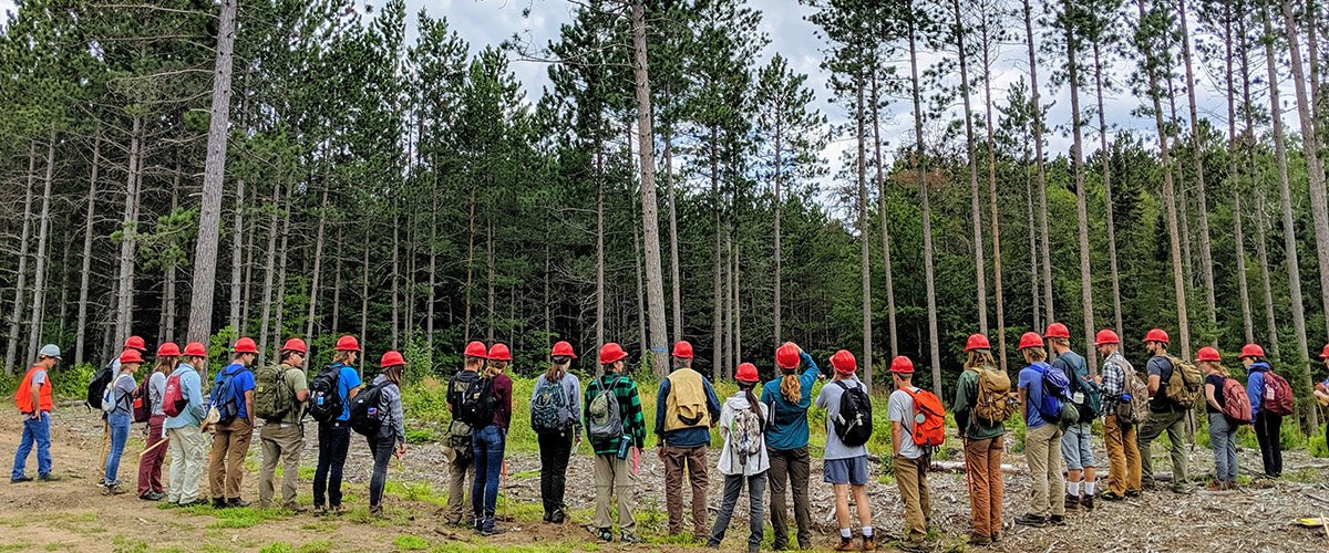 students wearing helmets lined up at the edge of a forest listening to an instructor